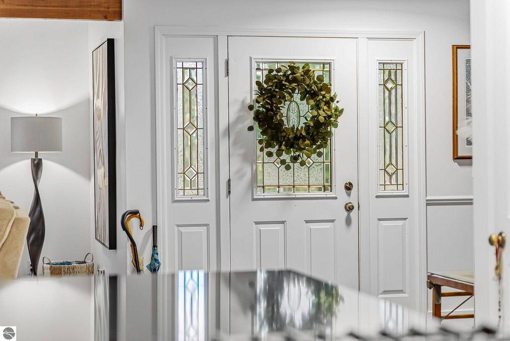 Front entryway of Lake Ann luxury home featuring a white door with decorative glass panels, a green wreath, modern lamp, and stylish decor, showcasing the inviting atmosphere of the property.