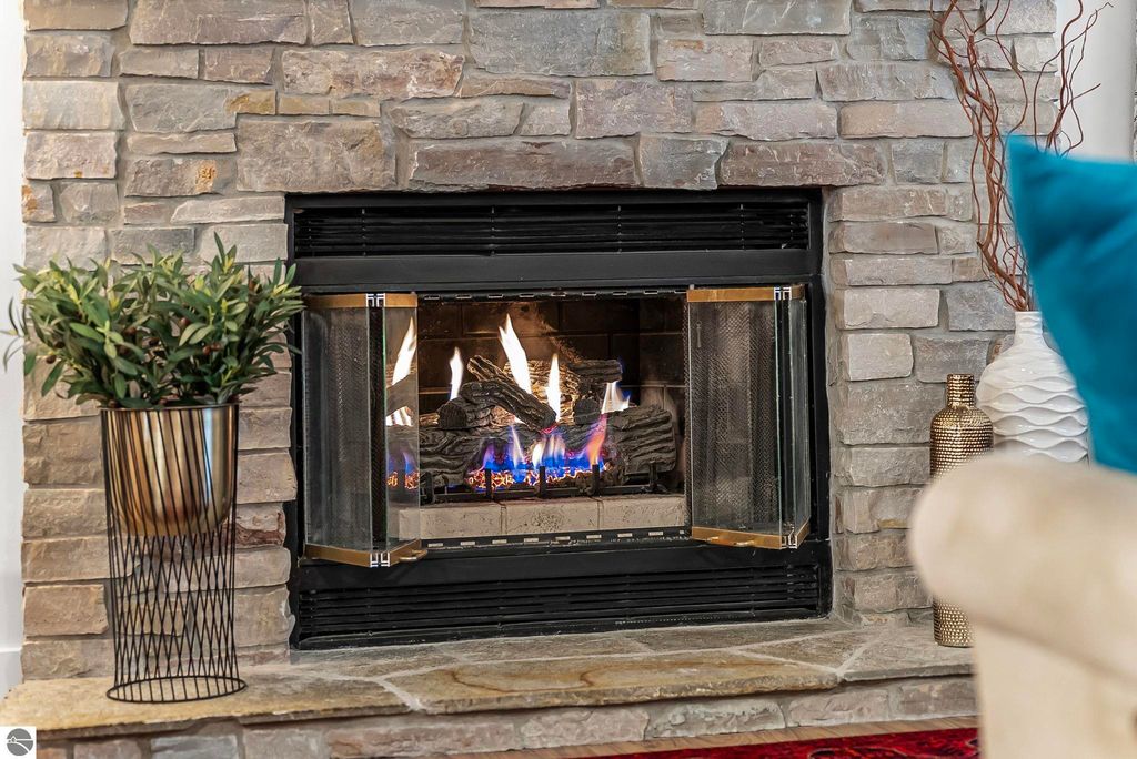 Cozy stone fireplace with flames, surrounded by modern decor elements including a potted plant, decorative vases, and a stylish armchair, in a luxurious Lake Ann home listing.