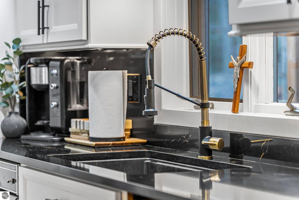 Modern kitchen with black quartz countertop, stylish faucet, coffee maker, paper towel holder, and decorative cross by the window, highlighting luxury features of the Lake Ann home.