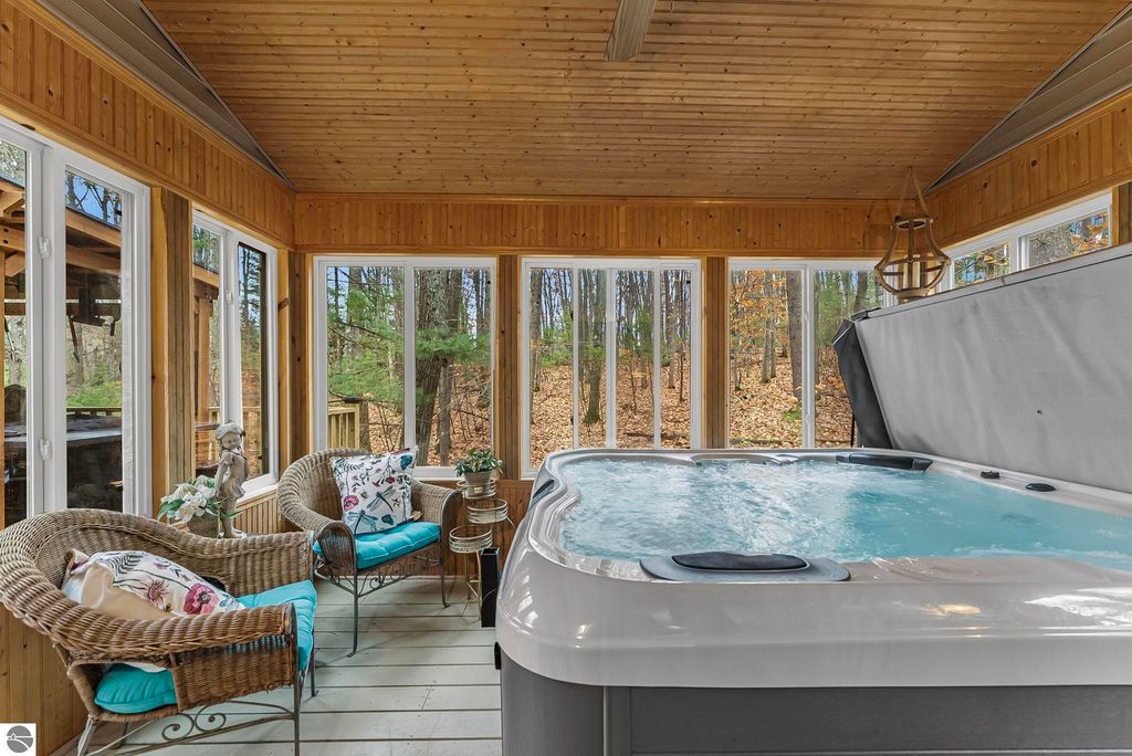 Hot tub in sunroom with wicker chairs and wooded view, showcasing luxury living in Lake Ann, MI home.