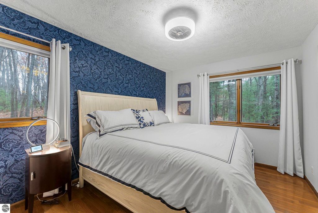 Modern bedroom interior with a large bed, floral blue wallpaper, wooden flooring, and windows showcasing a wooded view, emphasizing comfort and tranquility in the Lake Ann home for sale.