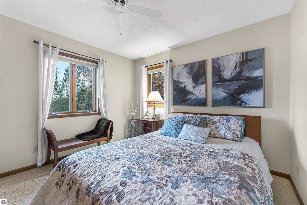 Cozy bedroom with a queen bed adorned in floral bedding, large windows with natural light, modern artwork on the walls, and a comfortable chair, showcasing the inviting interior of the luxury home at 5347 Birch Glen Road, Lake Ann, MI.