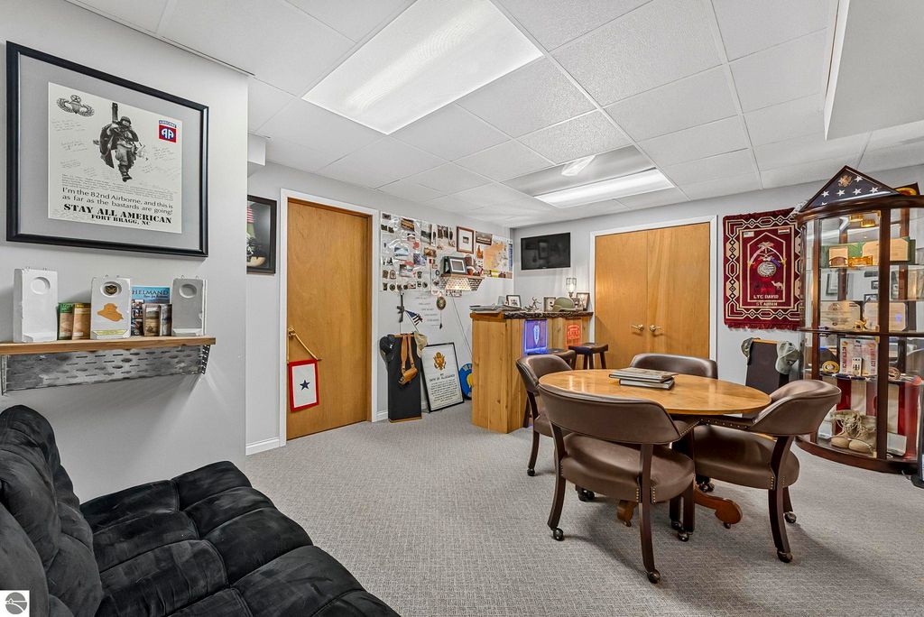 Cozy interior of a recreational room featuring a round wooden table with four chairs, a black couch, military-themed decor, and a display cabinet showcasing memorabilia, emphasizing the unique character of the Lake Ann home for sale.