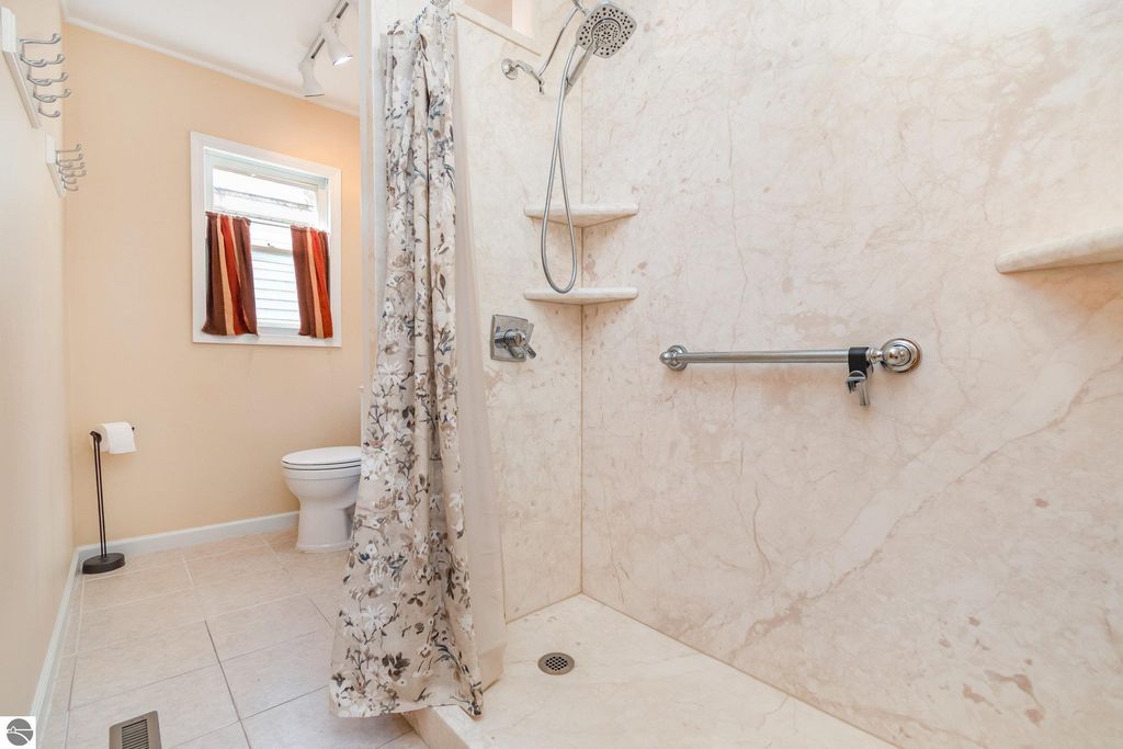 Modern bathroom with walk-in shower, floral curtain, toilet, and window with red and white curtains, featuring beige tile flooring and accessible design elements.