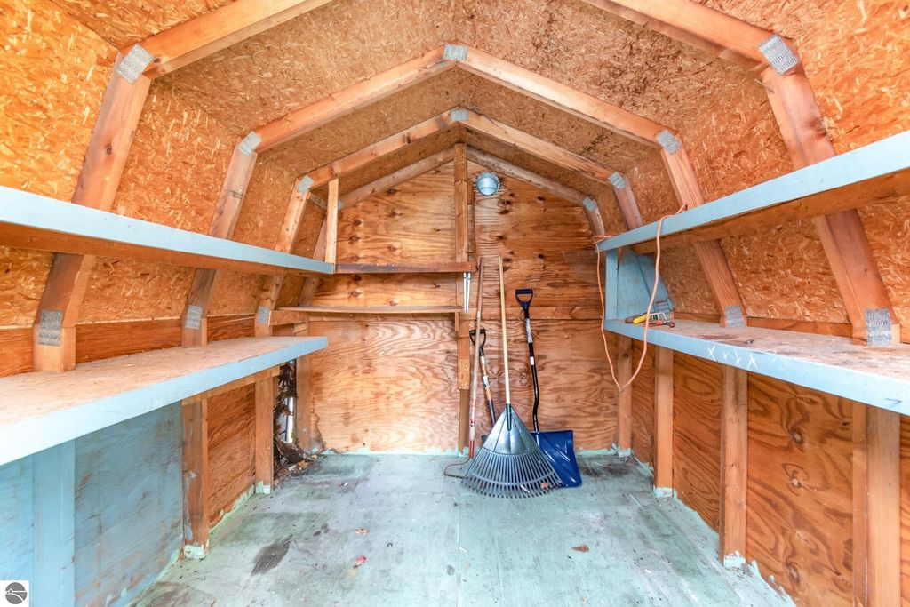 Interior of a storage shed featuring wooden walls, shelves, and gardening tools including a rake and a shovel, highlighting storage space in the backyard of the 2-bedroom, 1-bath home at 1435 E Broadway Street, Mt Pleasant, MI.