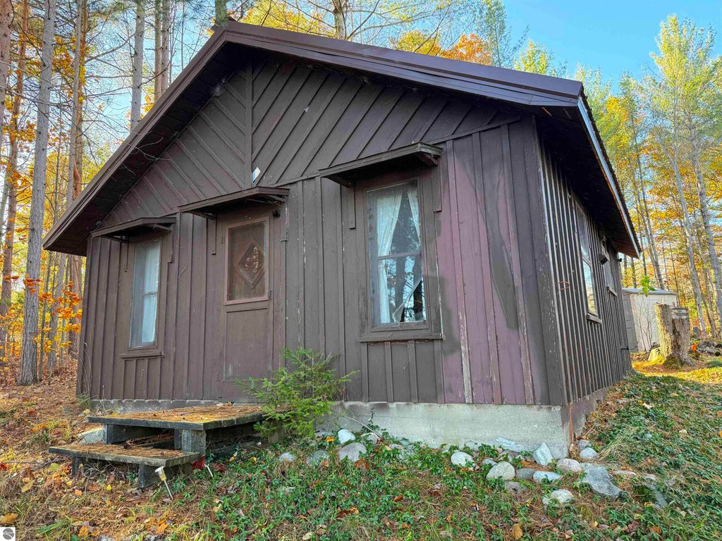 Older cabin on wooded lot near Pickerel Lake, featuring brown exterior, windows with white curtains, and steps leading to the entrance, surrounded by autumn foliage.