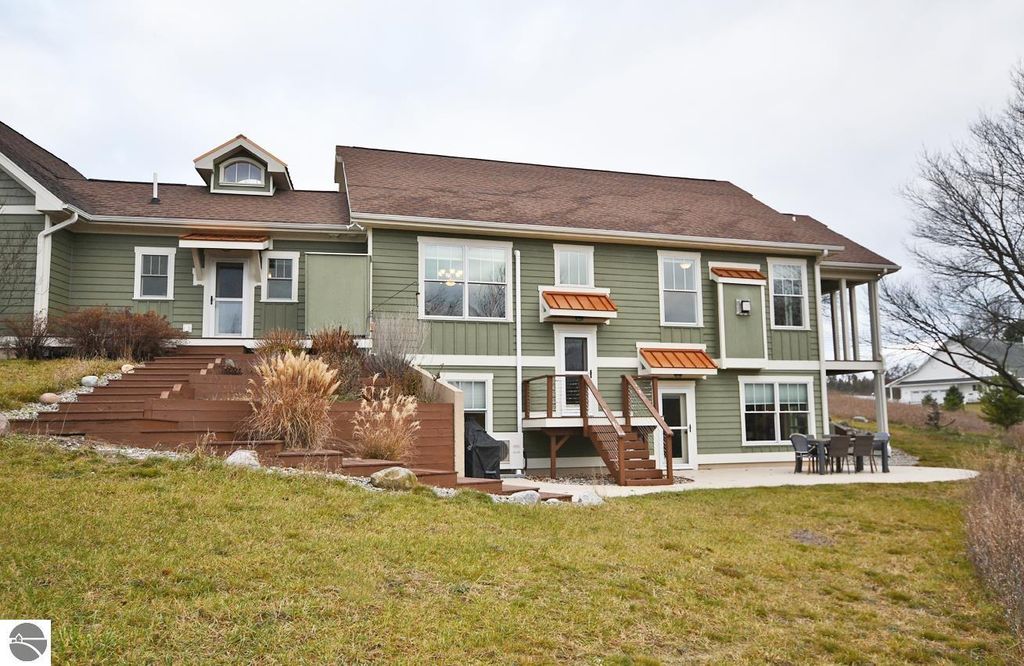 Exterior view of a green two-story home at 11007 S Rolling Meadows Drive, showcasing wooden stairs leading to a deck, surrounded by grassy land and landscaping, emphasizing a serene homestead lifestyle in Maple City, MI.
