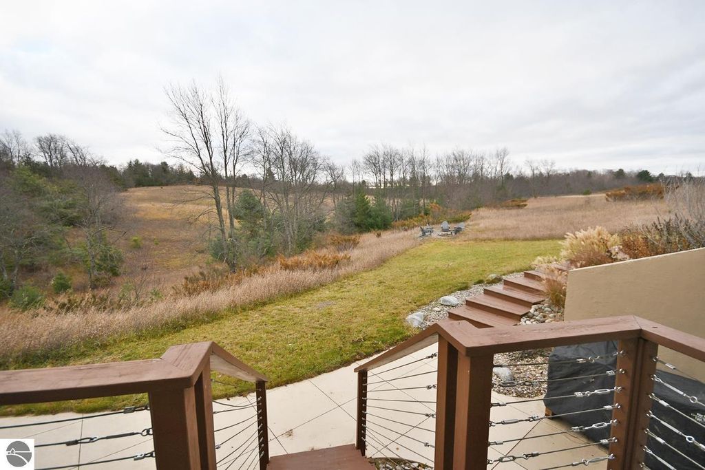 View from a wooden deck overlooking a spacious, grassy yard with rolling hills, trees, and a fire pit area, showcasing the serene landscape of the property at 11007 S Rolling Meadows Drive, Maple City, MI.