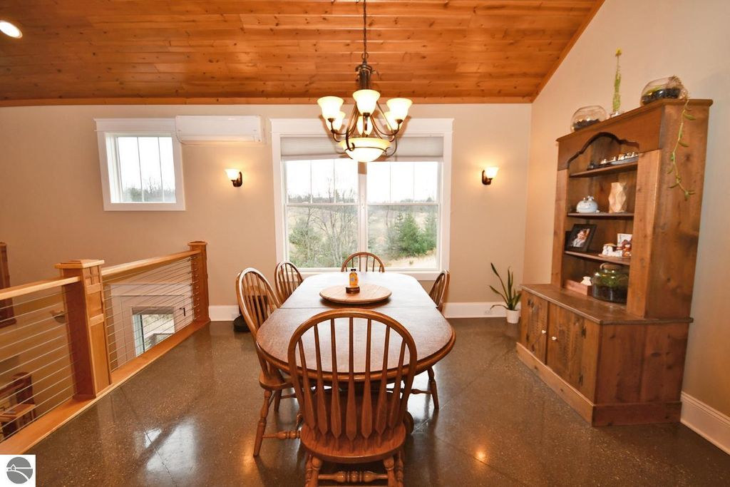 Dining area with wooden table and chairs, vaulted wood ceiling, large windows providing natural light, and a wooden cabinet, showcasing a cozy and inviting atmosphere in the home at 11007 S Rolling Meadows Drive, Maple City, MI.