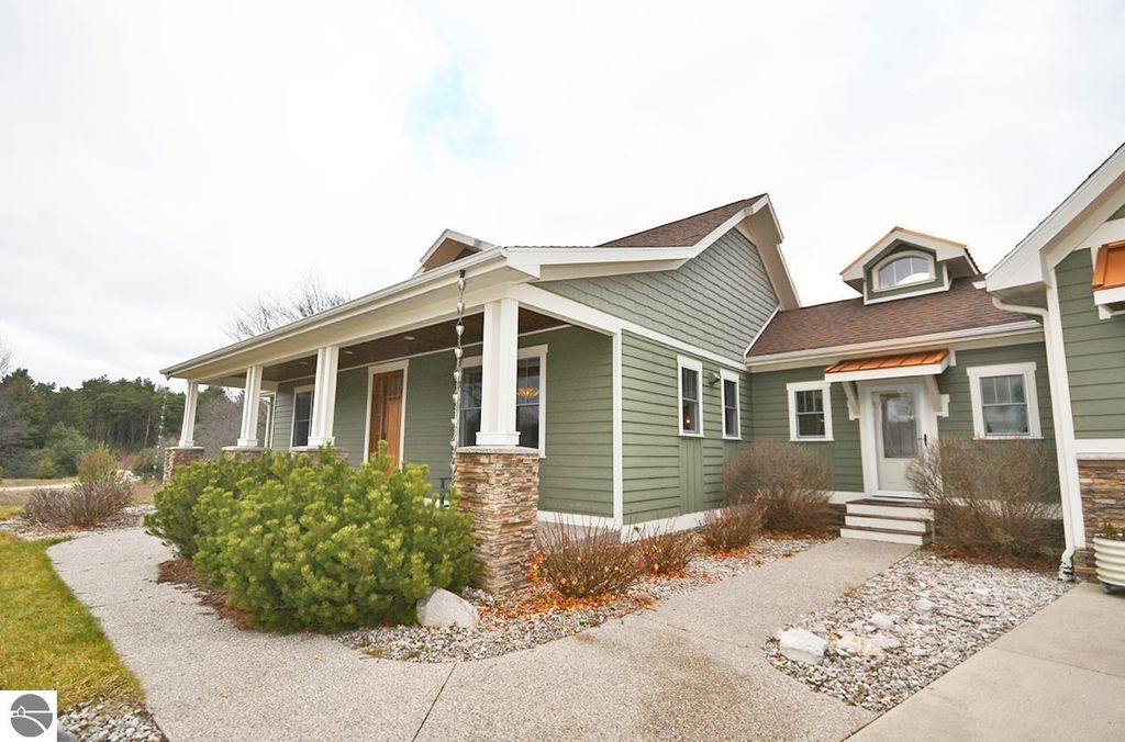 Exterior view of the home at 11007 S Rolling Meadows Drive, showcasing green siding, stone accents, and a welcoming porch, surrounded by landscaped pathways and natural greenery, emphasizing the property's serene homestead lifestyle on nearly 10 acres in Maple City, MI.