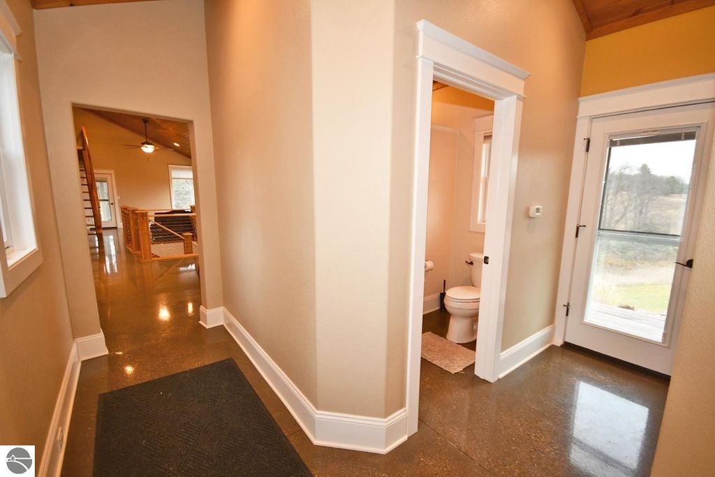 Interior hallway of a modern home featuring polished concrete flooring, a doorway leading to a bathroom with a toilet, and a large window providing natural light and a view of the surrounding landscape.