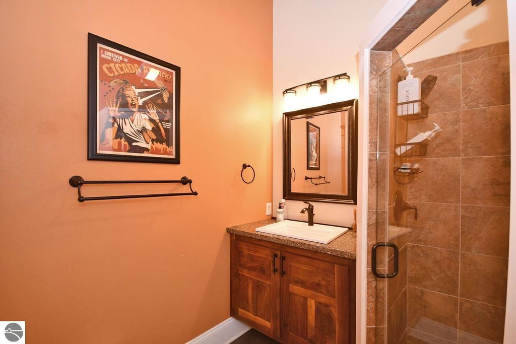 Bathroom interior featuring a wooden vanity with sink, framed mirror, and shower area, accented by warm orange walls and a decorative vintage-style poster.