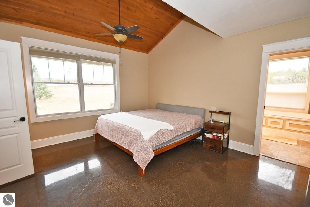 Modern bedroom with vaulted ceiling, large window showcasing outdoor view, hardwood floor, and minimalist decor, part of a four-bedroom home in Maple City, MI.