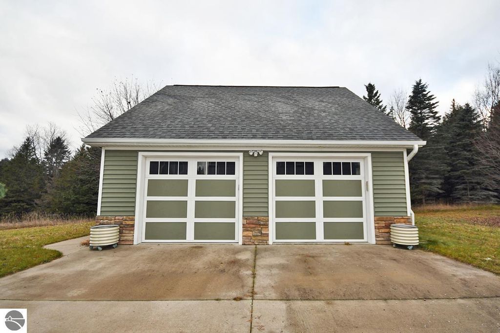 Detached garage with dual green doors and stone accents, set on a paved driveway surrounded by grassy landscape and trees, highlighting the property at 11007 S Rolling Meadows Drive, Maple City, MI.