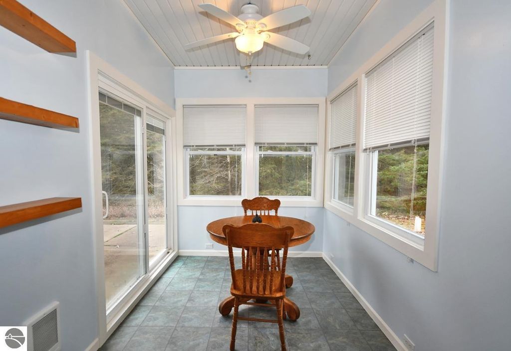 Bright sunroom with a wooden dining table and chairs, large windows showcasing wooded views, and a ceiling fan, part of a four-bedroom, three-bath home in Maple City, MI, emphasizing a serene lifestyle on nearly 10 acres.