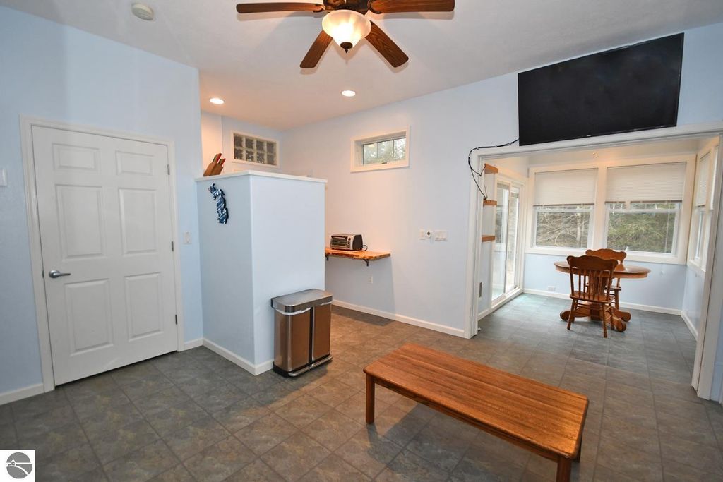 Interior view of a light blue room featuring a wooden bench, a dining table with chairs, a flat-screen TV mounted on the wall, and a door leading to an outdoor area, emphasizing the spacious layout and inviting atmosphere of the home at 11007 S Rolling Meadows Drive, Maple City, MI.