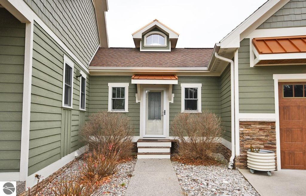 Front entrance of a modern four-bedroom home in Maple City, MI, featuring green siding, a gabled roof, and a welcoming porch with stone accents, surrounded by landscaped gravel and shrubs.