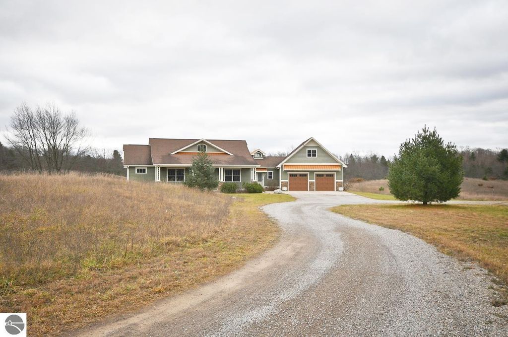 Stately four-bedroom home at 11007 S Rolling Meadows Drive, surrounded by rolling hills and meadows, featuring a gravel driveway, two-car garage, and landscaped yard, ideal for a homestead lifestyle in Maple City, MI.