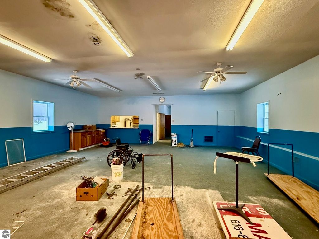 Interior view of a spacious room with blue and white walls, featuring a ceiling fan, large windows, and various items including a ladder, furniture, and tools, suitable for potential commercial use in Cadillac, MI.