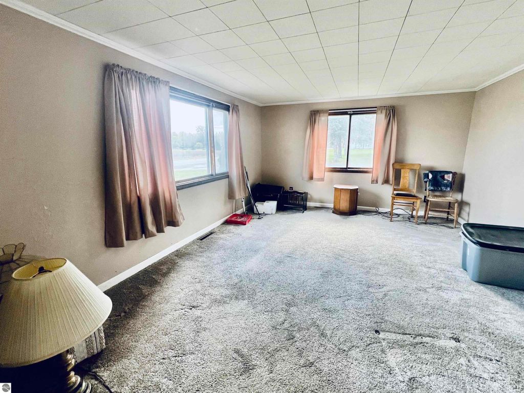 Living room interior of a three-bedroom home at 889 N Ferris Road, featuring large windows, beige walls, and carpet flooring, with minimal furniture including two chairs, a small table, and a lamp, highlighting the property's spaciousness and potential for personalization.