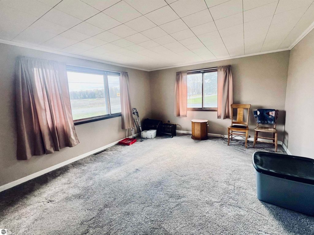 Spacious living room with large windows and natural light, featuring beige walls, light carpet, and minimal furniture including two chairs, a small table, and a storage bin, in a charming three-bedroom home at 889 N Ferris Road, Sumner, MI.
