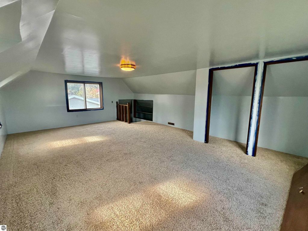 Spacious, carpeted upper room with sloped ceiling, large window, and wooden shelving, showcasing the potential for a cozy living space in the three-bedroom home at 889 N Ferris Road, Sumner, MI.