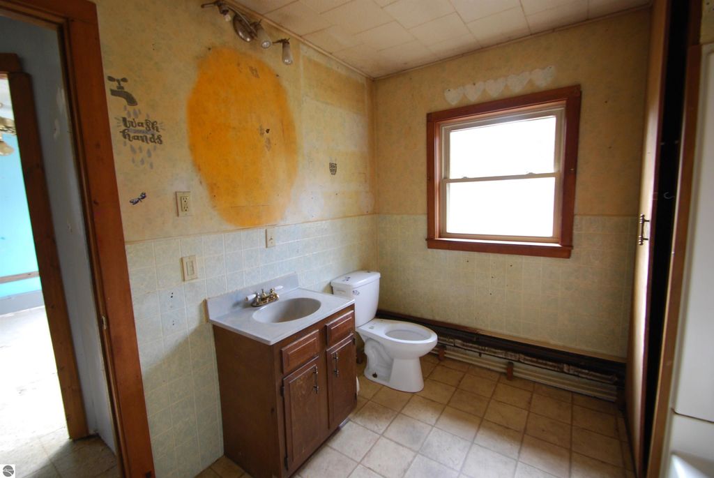 Bathroom interior featuring a sink with wooden cabinet, toilet, and window, showcasing potential for renovation in fixer-upper home at 1532 Rabbit Track Drive, Alger, MI.