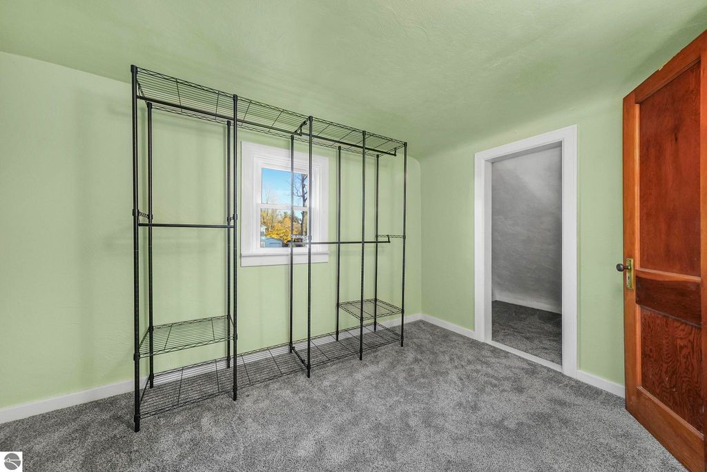 Updated bedroom with light green walls, gray carpet, and black wire shelving unit, featuring a window and door to a closet in a home at 310 E Euclid Street, McBain, MI.