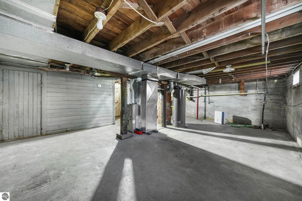 Basement interior of a home at 310 E Euclid Street, McBain, MI, featuring exposed wooden beams, concrete flooring, utility appliances, and a door leading to another room.