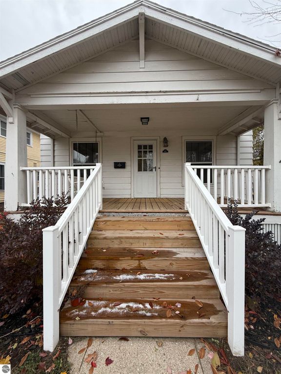 Front entrance of a residential property at 610 S Washington Street, Mt Pleasant, MI, featuring wooden steps, white railings, and a welcoming porch with a door and windows, surrounded by autumn foliage.