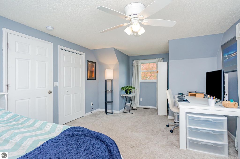 Updated bedroom with light blue walls, a ceiling fan, a workspace with a desk and computer, a cozy bed with patterned bedding, and a window with curtains, showcasing a bright and inviting atmosphere.