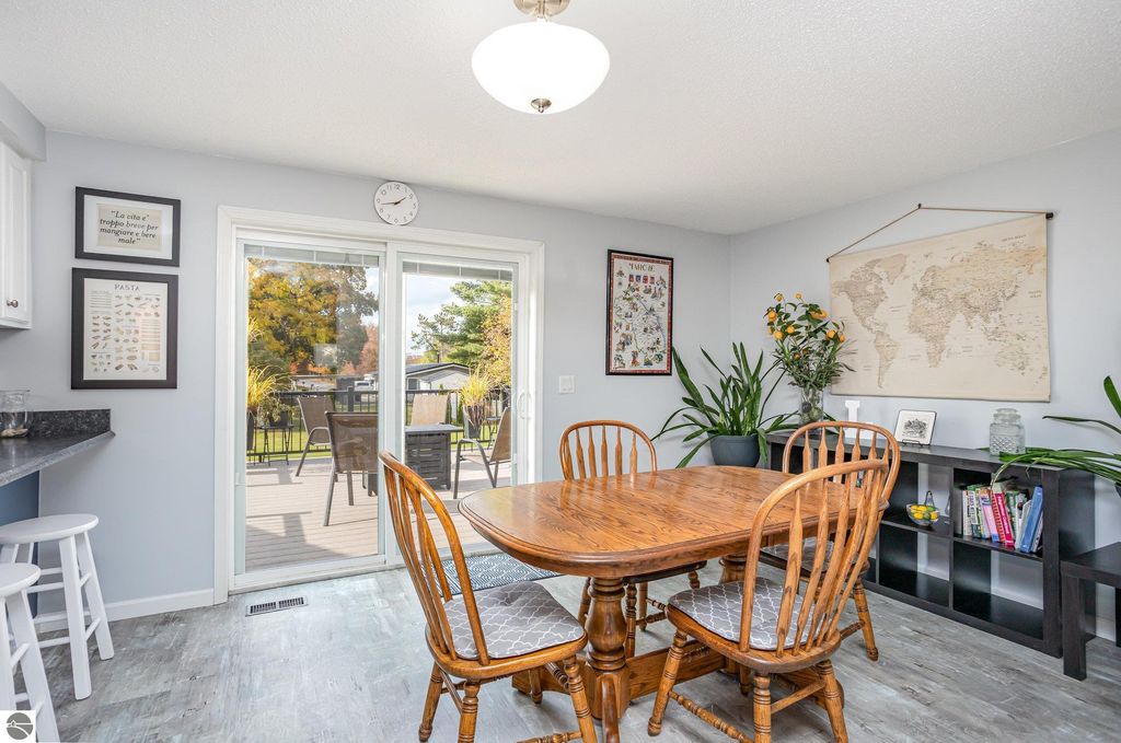 Dining area with wooden table and chairs, large sliding glass door leading to outdoor deck, wall decor including a world map and framed prints, plants adding greenery, modern kitchen setup with black countertops.