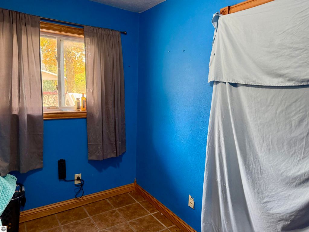 Interior view of a blue-painted bedroom with a window dressed in gray curtains, featuring a covered closet area and a tiled floor, showcasing the cozy atmosphere of the home at 325 Main Street, Breckenridge, MI.
