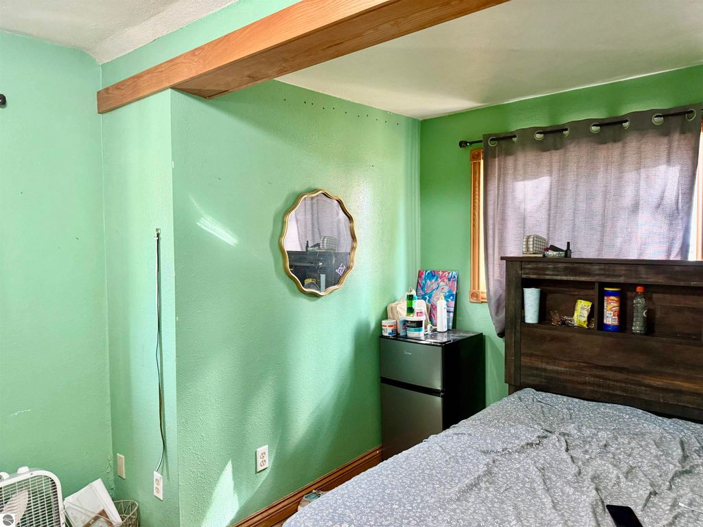 Cozy bedroom with green walls, a mirror, and a small fridge, featuring a bed with a patterned comforter and a window with gray curtains.