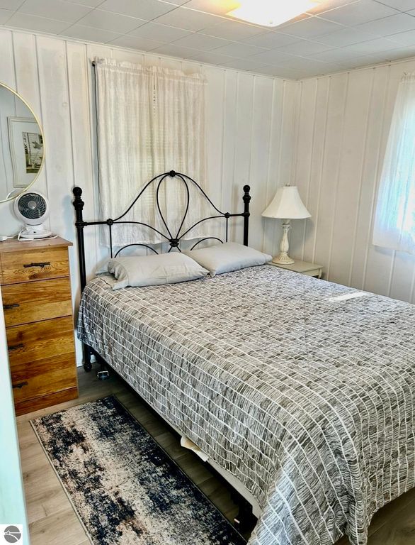 Cozy bedroom interior featuring a black metal bed frame with gray bedding, wooden dresser, bedside lamp, and natural light from windows, showcasing the charm of a cottage on Lake Missaukee.