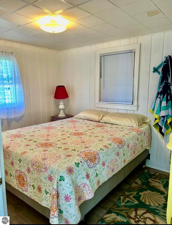 Cozy bedroom interior featuring a double bed with a floral quilt, a red bedside lamp, and a window with sheer curtains, highlighting the charm of the cottage on Lake Missaukee.