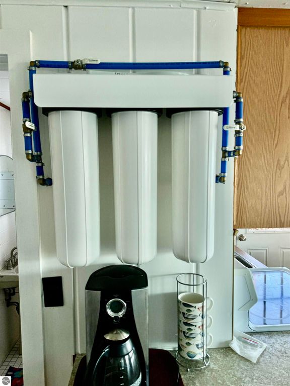 Water filtration system with three white filter cartridges mounted on a wall, featuring blue plumbing connections, alongside a coffee maker and a cup holder with patterned mugs in a kitchen setting.