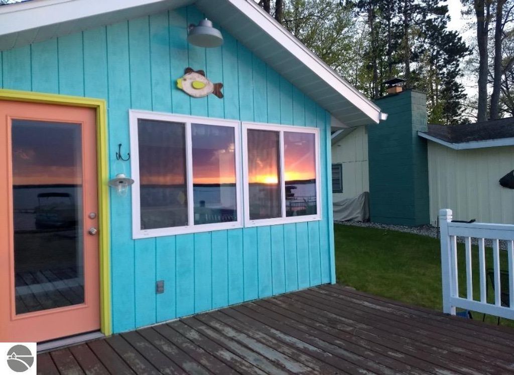 Cottage exterior on Lake Missaukee with turquoise siding, orange door, and sunset reflection in windows, showcasing lakefront charm and outdoor deck.