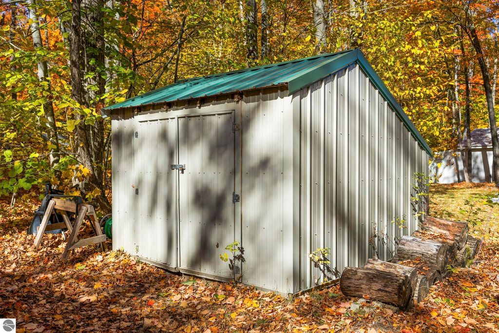 Metal storage shed surrounded by autumn foliage, featuring a green roof, positioned near logs and outdoor tools, reflecting the serene setting of the Blue Lake Heights community in Kalkaska, MI.