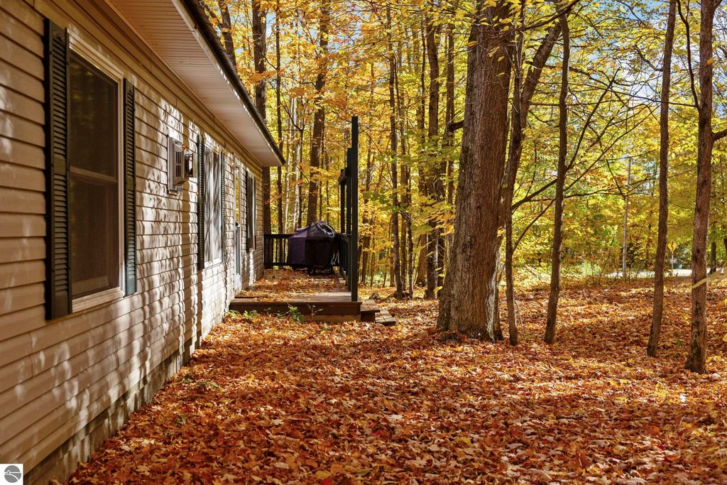 Side view of a home at 7149 W Blue Lake Road, surrounded by autumn foliage and fallen leaves, featuring a wooden deck and natural wooded area, emphasizing the serene outdoor setting in the Blue Lake Heights community.