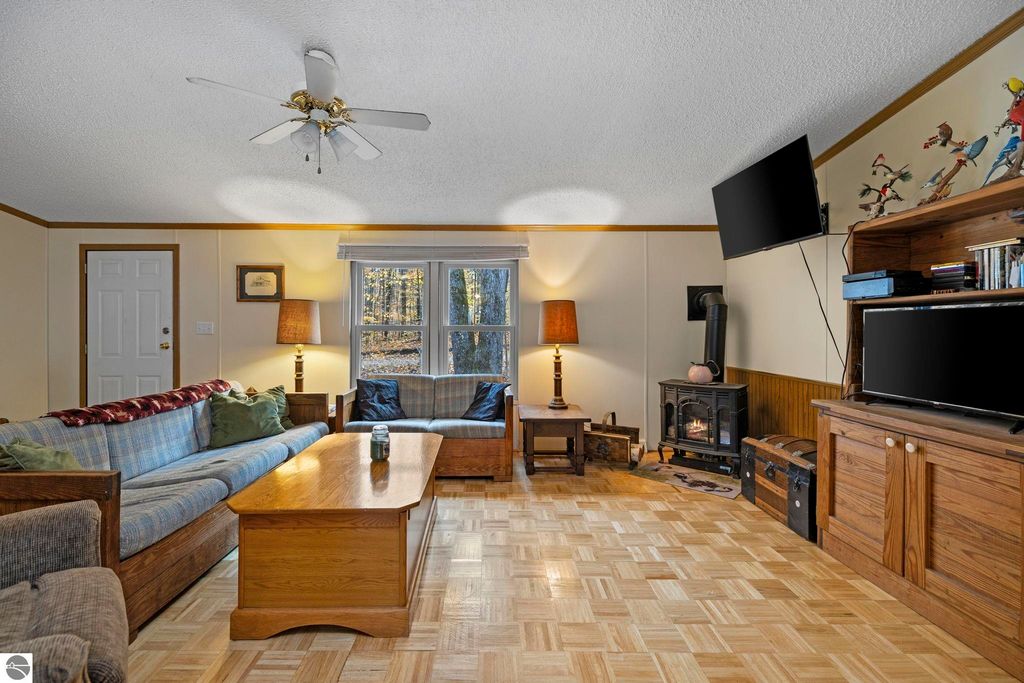 Cozy living room featuring a blue couch, wooden coffee table, and a wood-burning stove, with natural light streaming through large windows overlooking a wooded area, enhancing the inviting atmosphere of the home at 7149 W Blue Lake Road, Kalkaska, MI.