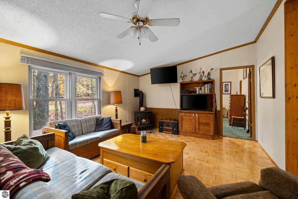 Inviting living room with cozy seating, wooden furniture, large windows showcasing autumn scenery, and a TV stand, highlighting the comfort and charm of the home at 7149 W Blue Lake Road, Kalkaska, MI.