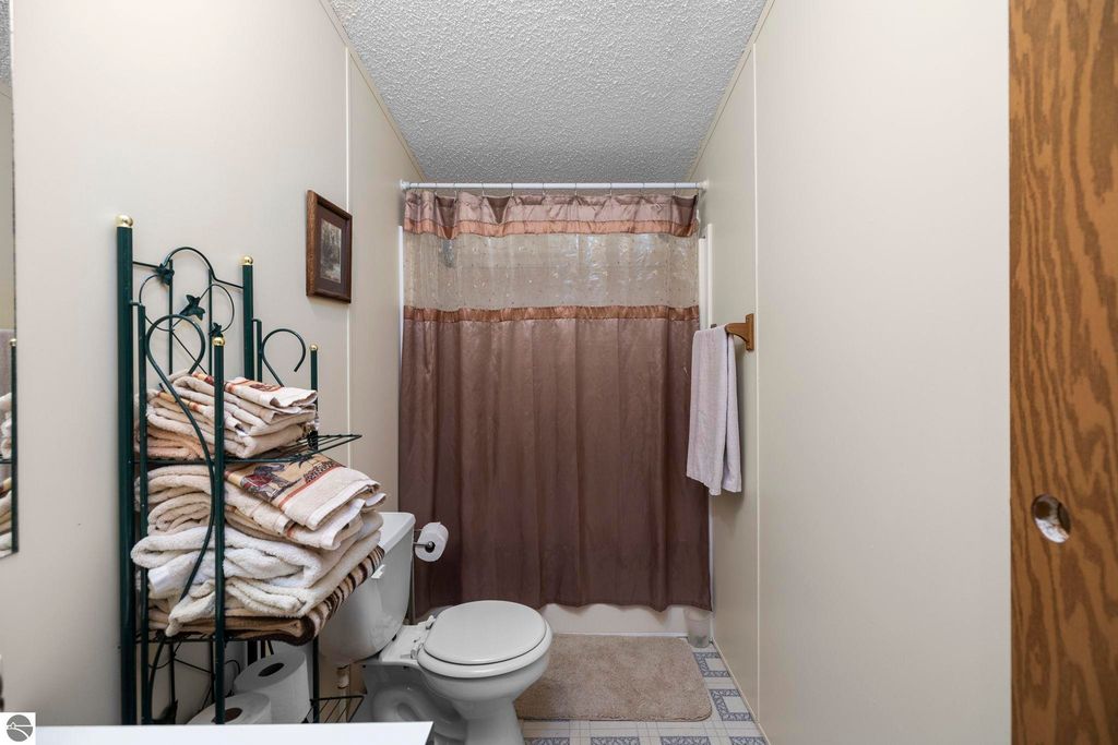 Bathroom interior featuring a toilet, shower with a brown curtain, stacked towels on a decorative rack, and light-colored walls, showcasing the home's comfort and functionality.
