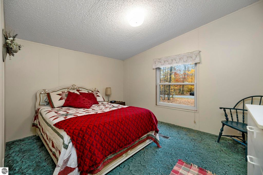 Cozy bedroom featuring a red and white quilted bedspread, window with lace curtains, and a view of autumn foliage outside, highlighting the inviting atmosphere of the home at 7149 W Blue Lake Road, NE, Kalkaska, MI.