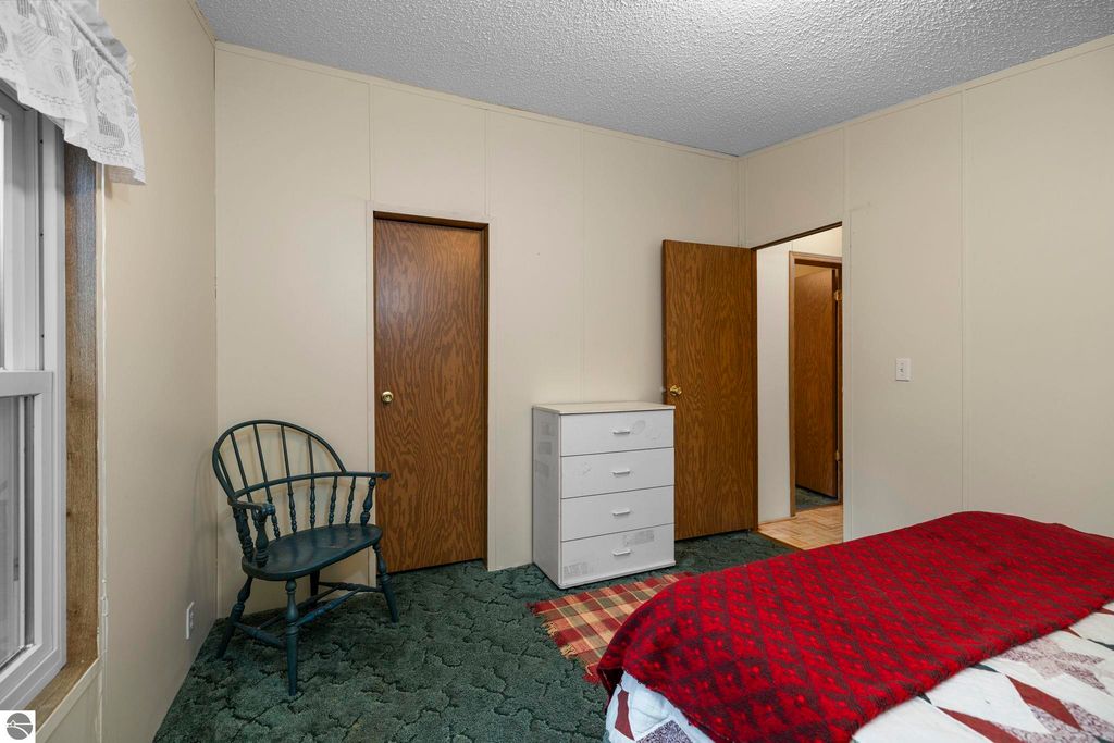 Interior view of a cozy bedroom featuring a red quilted bedspread, a white dresser, a green upholstered chair, and two wooden doors leading to adjacent rooms, emphasizing the home's inviting atmosphere.