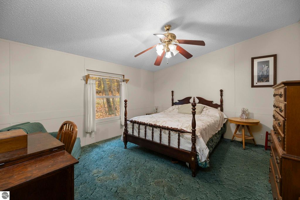 Cozy bedroom featuring a wooden bed with a floral bedspread, a vintage dresser, a desk, and a window with natural light illuminating the green carpet and forest view outside.