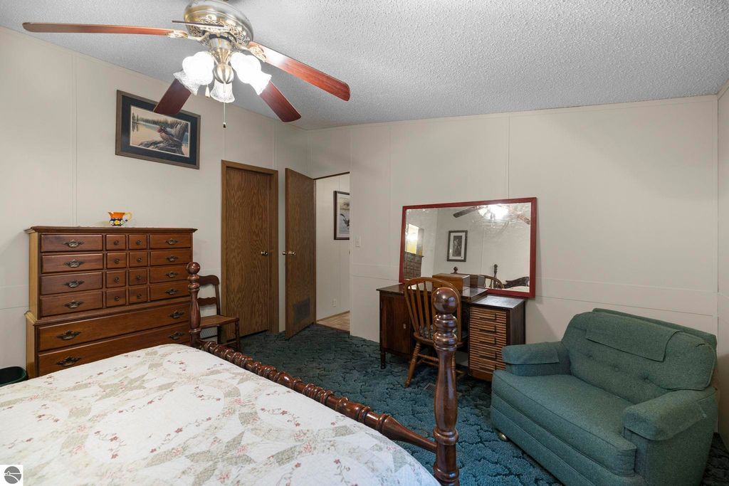 Cozy bedroom featuring a wooden dresser, a green armchair, and a mirror on a desk, with a ceiling fan and framed artwork on the wall, highlighting the inviting atmosphere of the home at 7149 W Blue Lake Road in Kalkaska, MI.