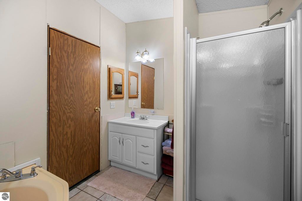 Bathroom interior with white vanity, sink, and mirrors, featuring a shower stall and wooden door, showcasing a cozy and functional space in a 3-bedroom home for sale in Kalkaska, MI.