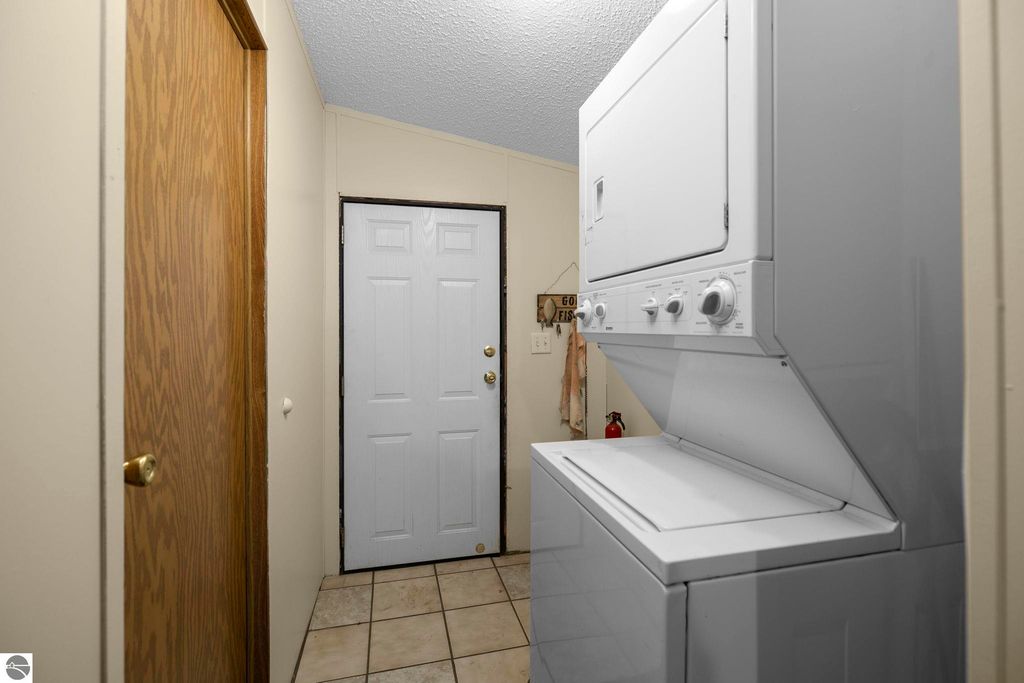 Laundry area with stacked washer and dryer, white door leading outside, and wooden door to a closet, tiled floor, and neutral walls.