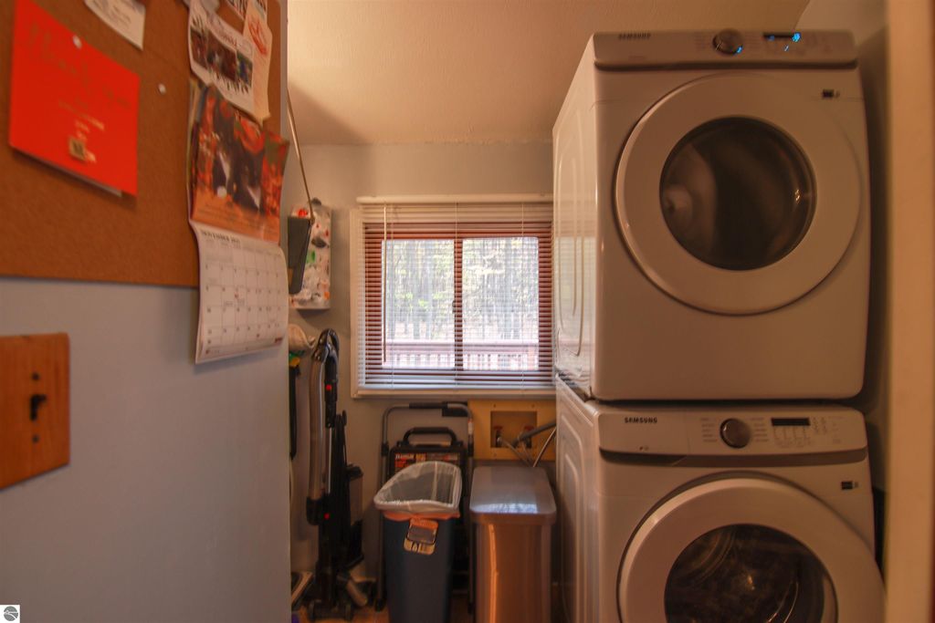 Laundry area featuring stacked Samsung washer and dryer, window with blinds, corkboard, calendar, and storage items in a cozy home setting.